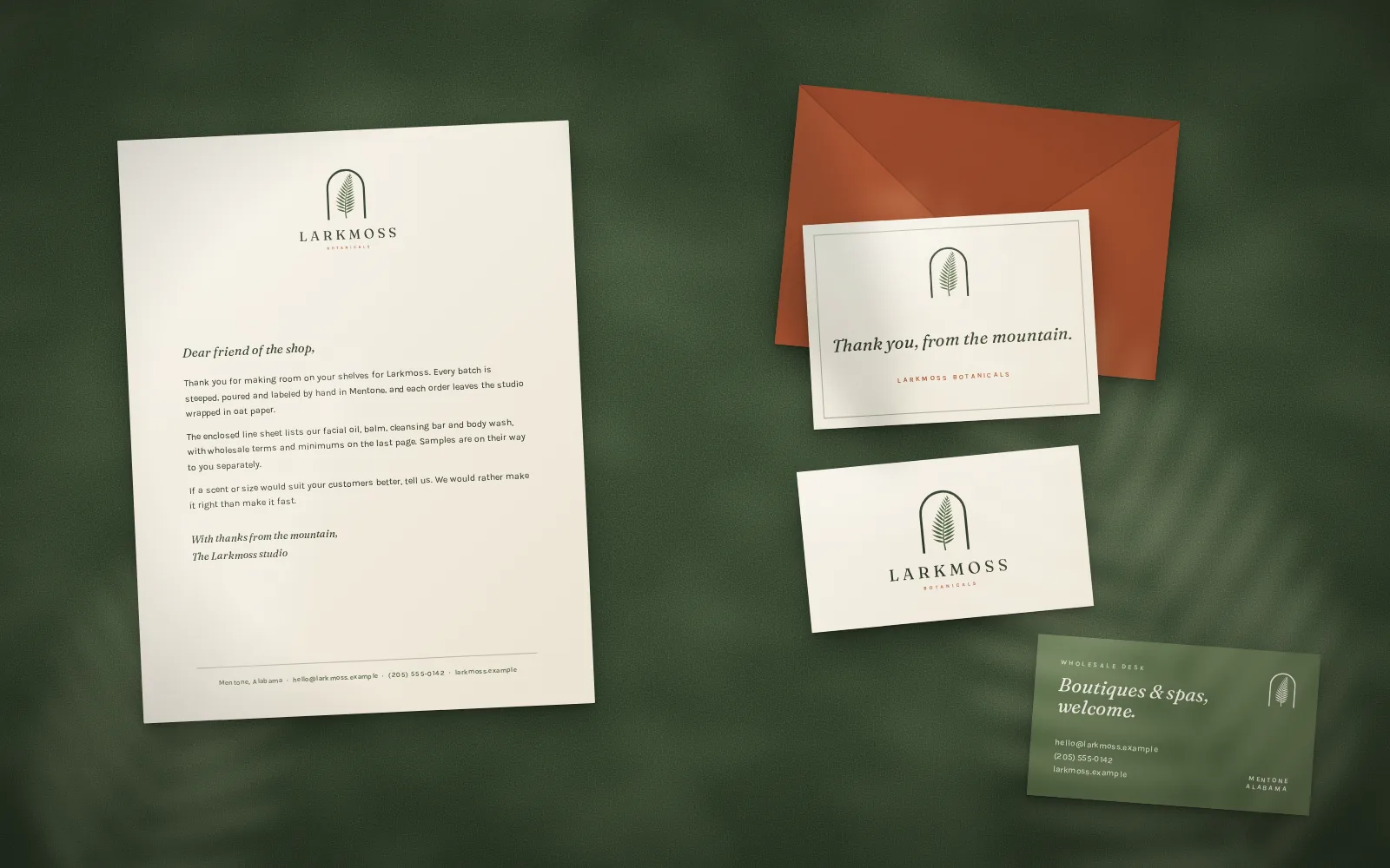 Larkmoss letterhead, a notecard on a clay envelope and two business cards laid out on a deep green felt surface.